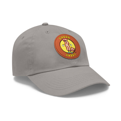 'Dad hat' style Grey coloured baseball cap with a round, brown leather patch sewn on the front. Printed on the leather patch is a yellow circular graphic logo of the Bacon Buddy character, a cartoon style strip of bacon with arms, legs and sunglasses. Curved yellow text of Bacon Buddy around the logo. Photographed is front / side view 