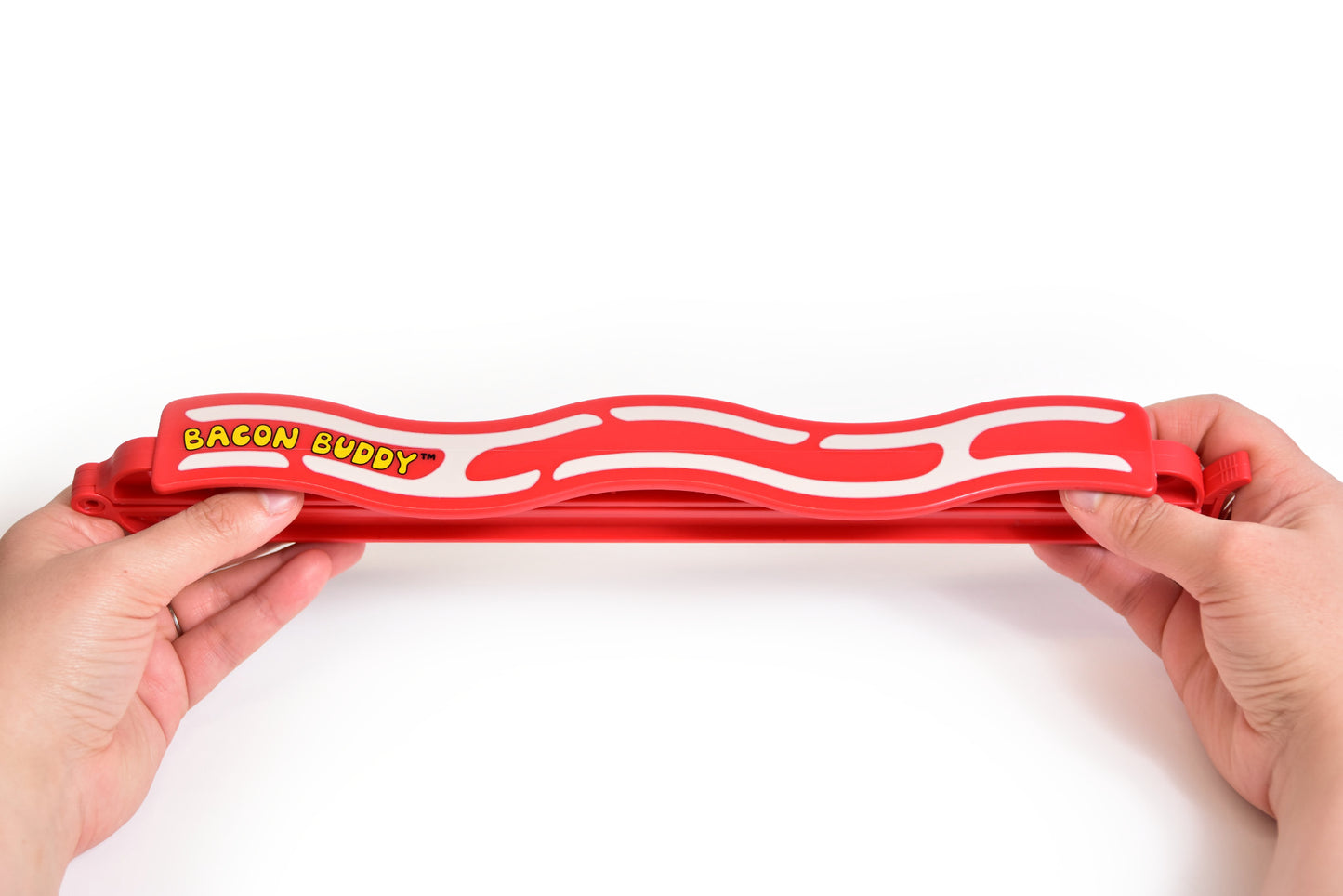 The Bacon Buddy clip, a 13 inch (35cm) long, red wavy bacon shaped plastic food packaging clip. Has printed wavy text in yellow saying 'Bacon Buddy' and wavy cream strips to look like bacon. Clip being held at both ends with two hands, photographed on a plain bright white background