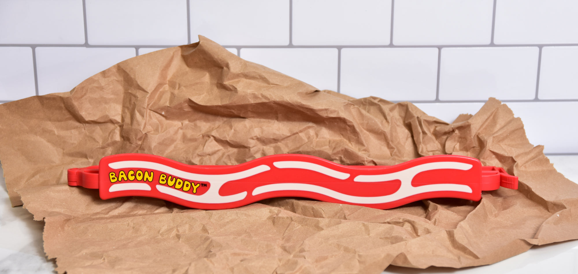 The Bacon Buddy clip, a 13 inch (35cm) long, red wavy bacon shaped plastic food packaging clip. Has printed wavy text in yellow saying 'Bacon Buddy' and wavy cream strips to look like bacon. Clip photographed on down paper on a kitchen counter top