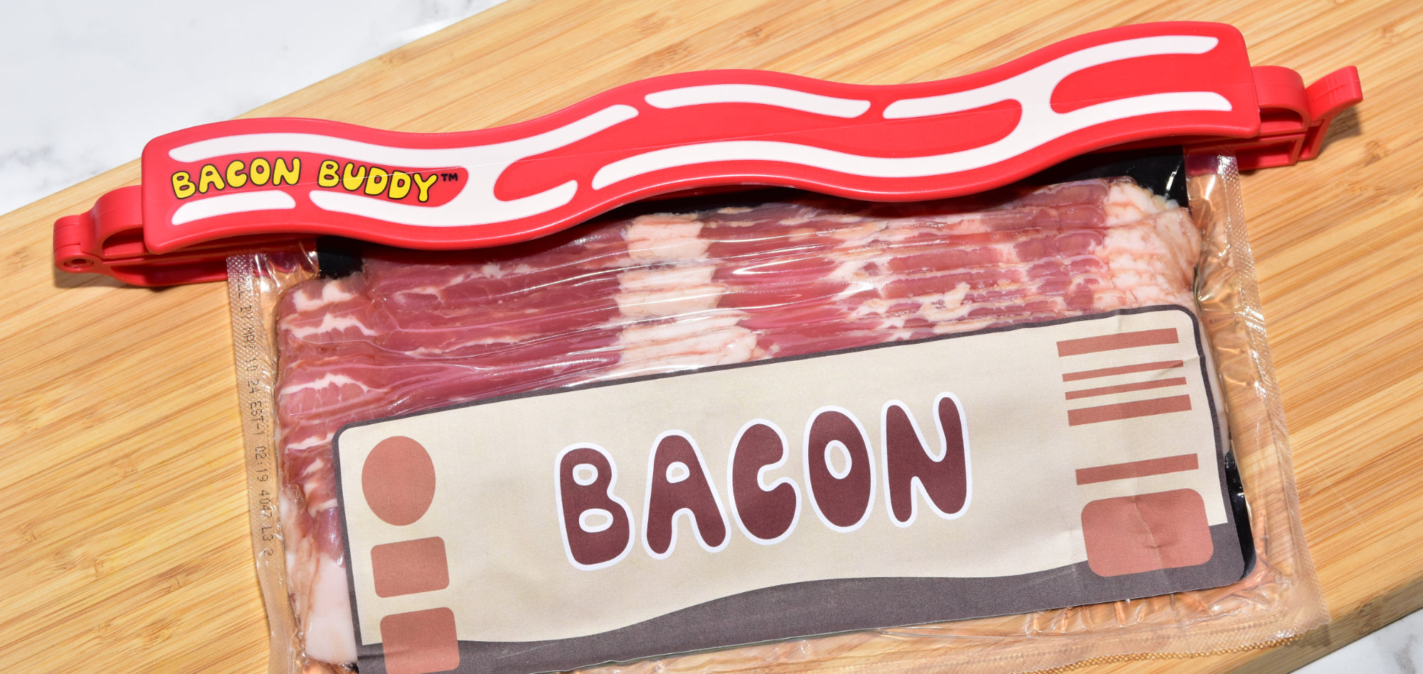 The Bacon Buddy clip, a 13 inch (35cm) long, red wavy bacon shaped plastic food packaging clip. Has printed wavy text in yellow saying 'Bacon Buddy' and wavy cream strips to look like bacon. Clip photographed closing a package of bacon on a kitchen counter top