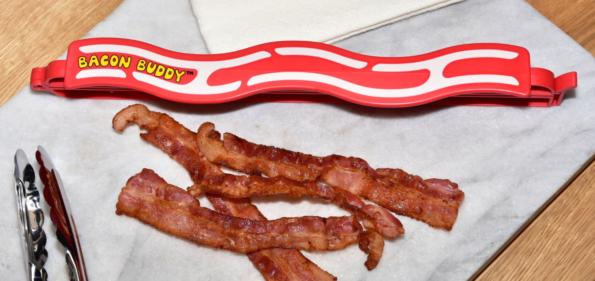 The Bacon Buddy clip, a 13 inch (35cm) long, red wavy bacon shaped plastic food packaging clip. Has printed wavy text in yellow saying 'Bacon Buddy' and wavy cream strips to look like bacon. Clip photographed next to cooked bacon and tongs on a cutting board